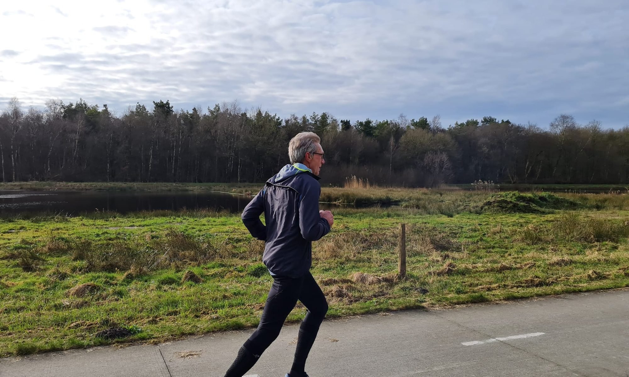 Oudere man met grijs haar jogt langs een ven onder bewolkte hemel